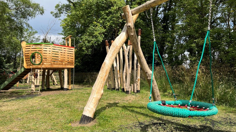 A large swing supported by wooden posts in the foreground, with a wooden boat-shaped climbing frame in the background.